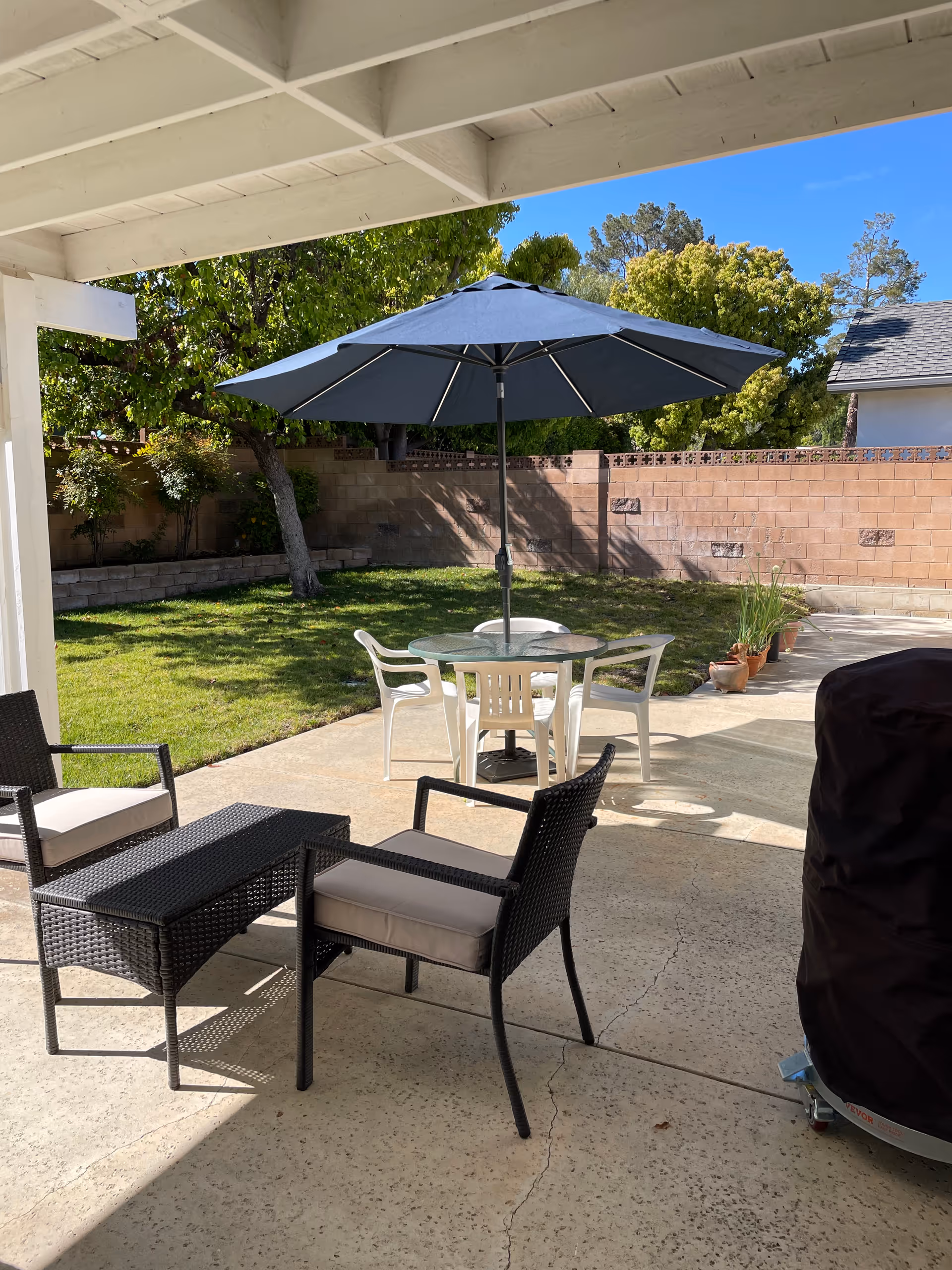 Outdoor patio area with two black wicker chairs with cushions and a matching table under a covered porch. In the background, there is a round glass table with four white plastic chairs and a large blue umbrella. The patio opens to a grassy yard with trees and a brick wall fence under a clear blue sky.