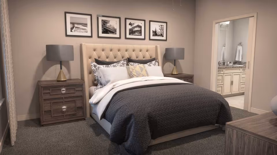 A cozy bedroom featuring a large bed with a tufted beige headboard, dark gray bedding, and multiple pillows. On each side of the bed are matching wooden nightstands with modern table lamps. Above the bed are four black-and-white framed photographs. To the right, an open doorway reveals a bathroom with a vanity and towels.