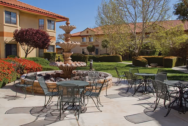 Sunny courtyard with a tiered fountain, metal patio tables and chairs, lawns and Mediterranean-style buildings with red tile roofs.