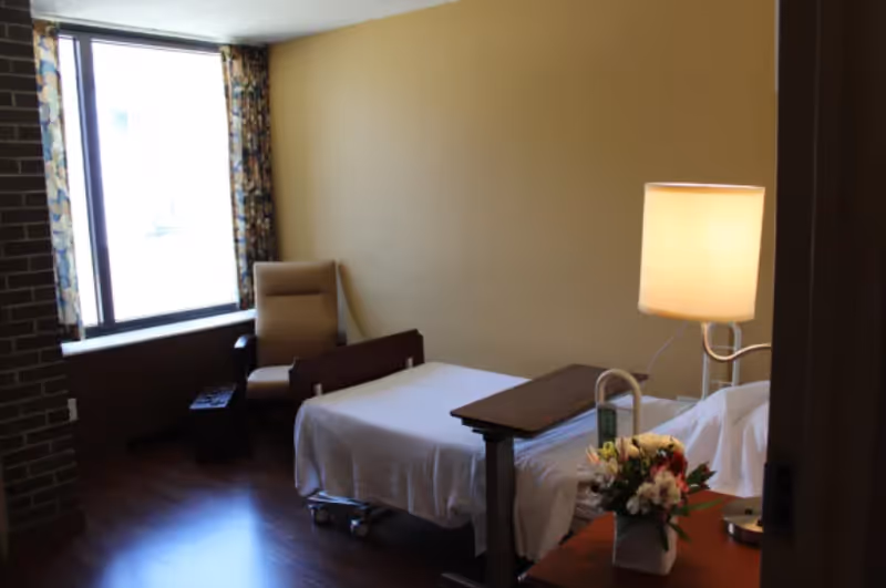 A small, simple bedroom with a single hospital-style bed covered with white sheets. Next to the bed is a wooden bedside table with a lamp and a vase of flowers. There is a beige armchair near a large window with patterned curtains, and the floor is wooden.