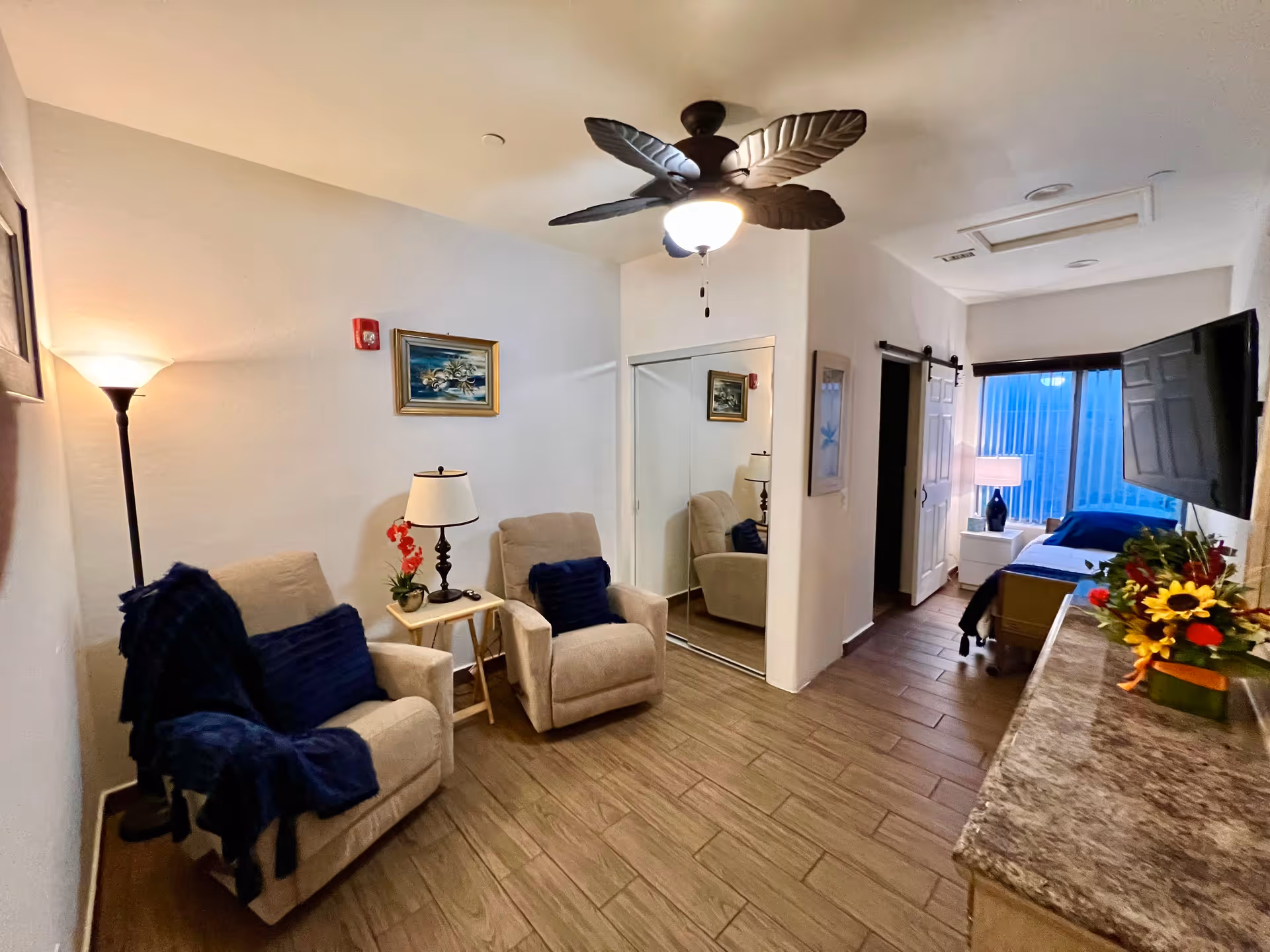 A cozy interior room with two beige armchairs with dark blue cushions and a throw blanket, a small side table with a lamp and a red flower, a ceiling fan with leaf-shaped blades, a mirrored closet door, a bed with blue bedding near a window with vertical blinds, a wall-mounted TV, and a dresser with a flower arrangement.