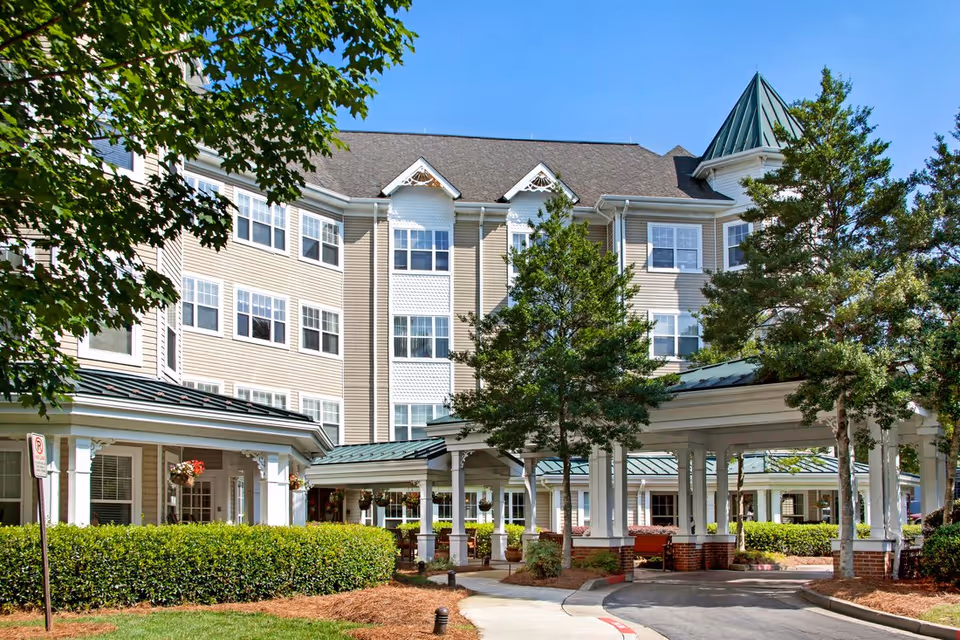 Multi-story senior living building with a covered porte-cochère entrance, landscaped shrubs, and trees under a clear blue sky.