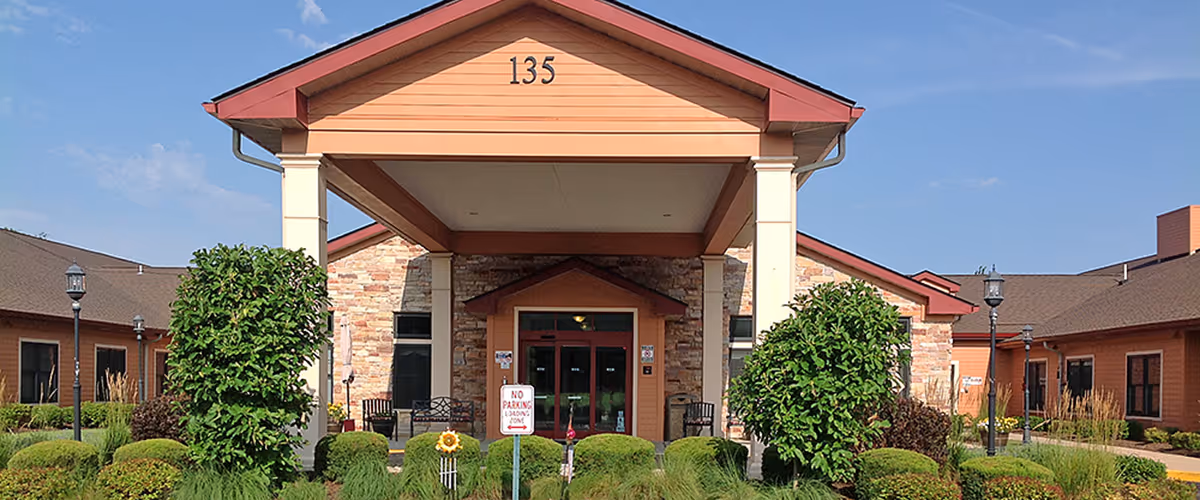 Front exterior view of a single-story building with a covered entrance supported by two white columns. The building has a stone facade around the entrance and orange siding with a brown roof. The number 135 is displayed at the top of the entrance. There are neatly trimmed bushes and greenery in front of the building, along with a 'No Parking Loading Zone' sign.