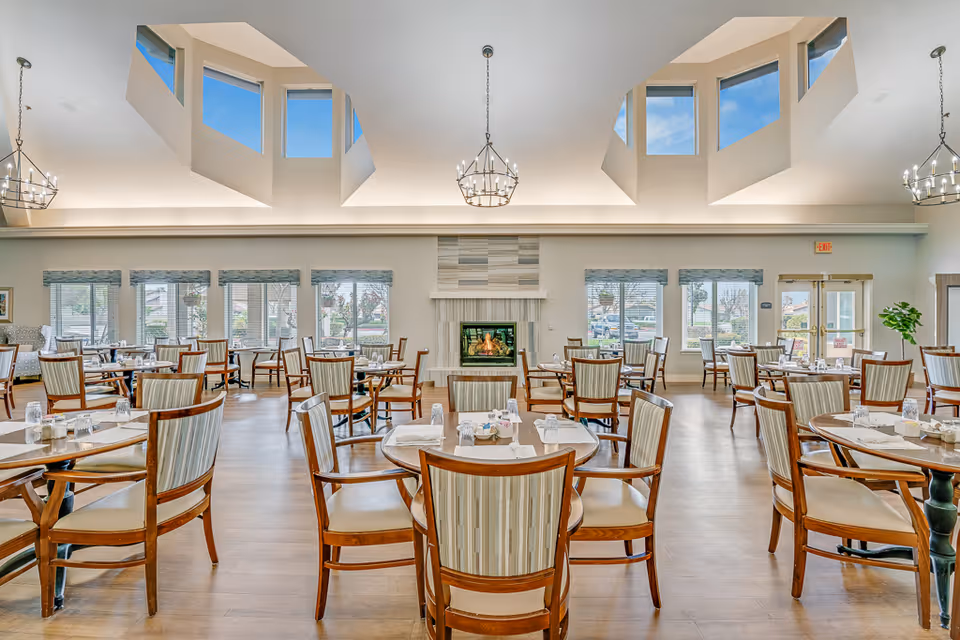 A spacious dining room with multiple round tables and wooden chairs with striped upholstery. The room features large windows with blinds, a fireplace in the center of the far wall, and modern chandeliers hanging from a high ceiling with skylights. The floor is wooden, and the room is well-lit with natural light.
