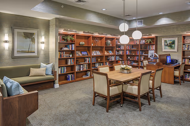 A cozy senior living facility common area with a wooden table and six upholstered chairs in the center. Behind the table are wooden bookshelves filled with books and decorative items. To the left, there is a cushioned bench with pillows under a framed painting. The room is softly lit with wall sconces and three round pendant lights hanging from the ceiling. A desk with a computer is visible in the back right corner.