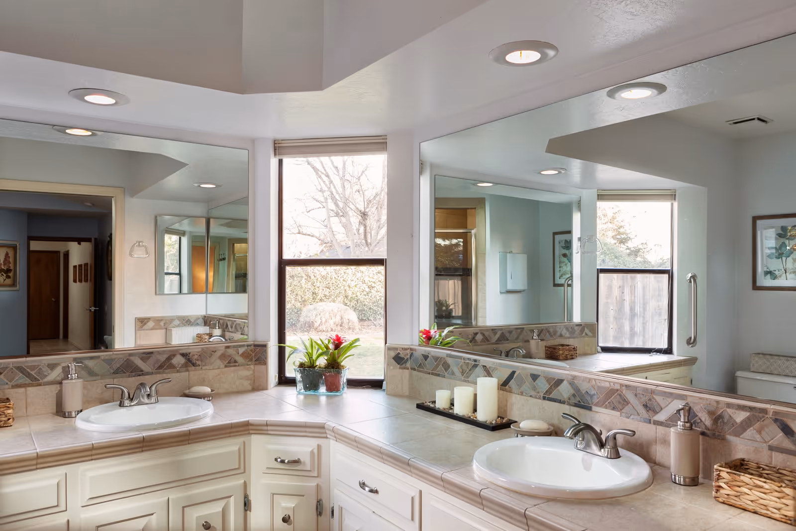 Bright bathroom featuring a corner vanity with two sinks, wide mirrors, tiled countertops, and a window with potted plants.
