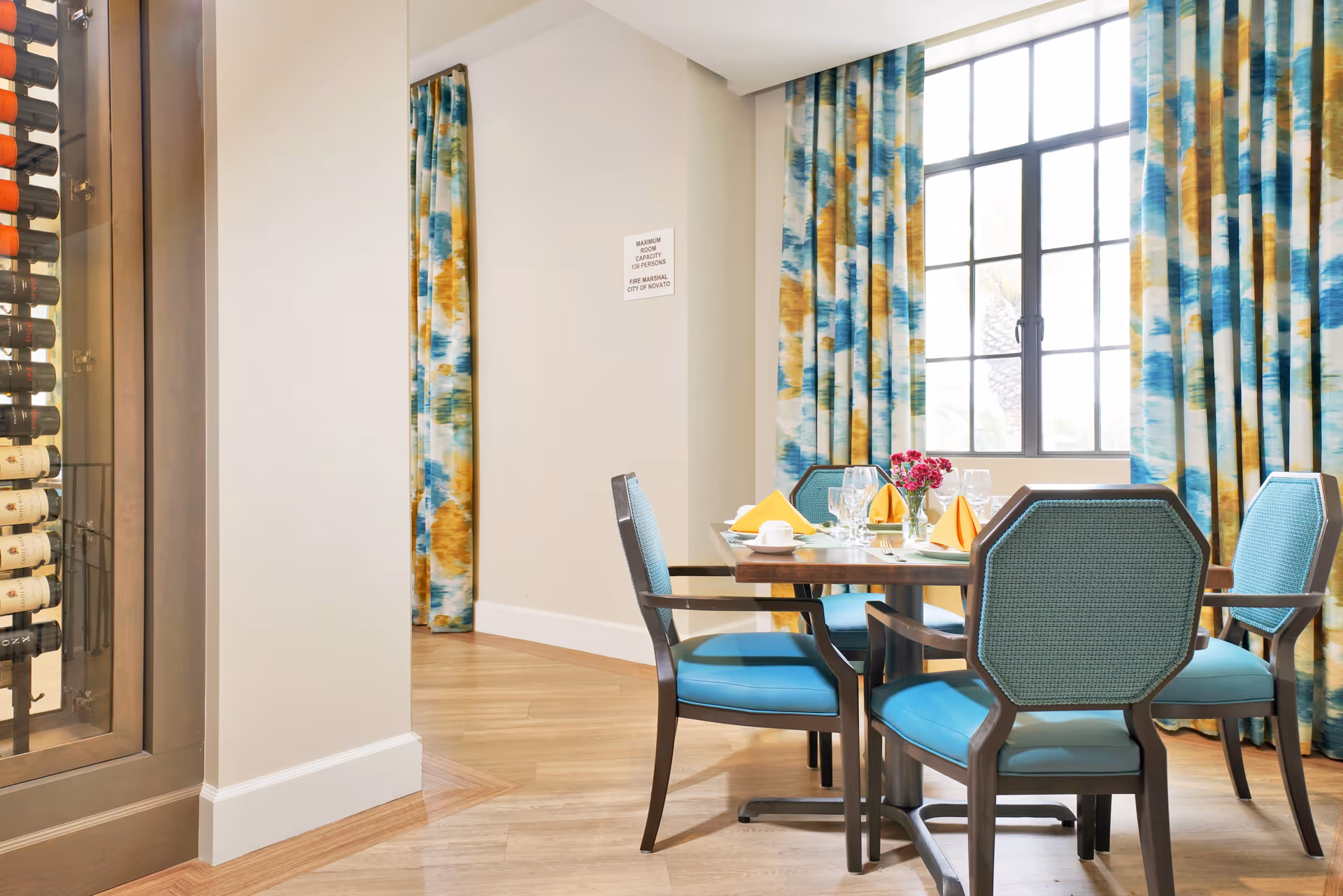 A dining area with a wooden table set for four people, featuring blue cushioned chairs, yellow folded napkins, glassware, and a small vase with pink flowers. The room has large windows with colorful blue, yellow, and white curtains, and a wine rack is visible on the left side.