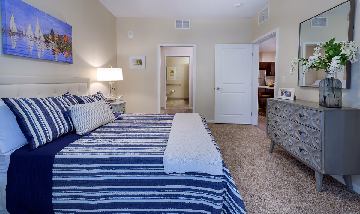 A neatly made bed with blue and white striped bedding in a bedroom. To the left of the bed is a nightstand with a lamp and a small clock. On the wall above the bed is a painting of sailboats on water. The room has beige walls and carpeted floor. There is a dresser with a decorative vase of flowers and a framed photo on the right side. Two open doorways lead to a bathroom and another room with a kitchen visible in the background.