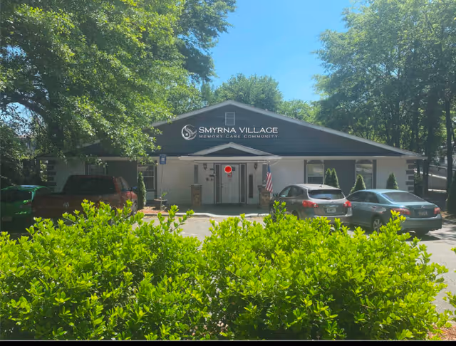 Front exterior view of Smyrna Village Memory Care Community building with a parking area in front, several cars parked, green bushes in the foreground, and large trees surrounding the building under a clear blue sky.