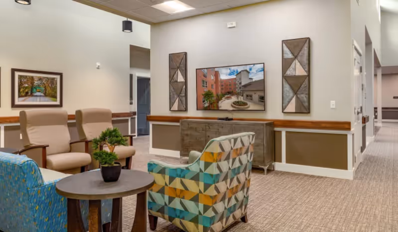 A senior living community lounge area with comfortable seating including two beige armchairs and two patterned armchairs around a small round table with a potted plant. A flat-screen TV is mounted on the wall above a wooden console, flanked by two decorative wall panels. The room has neutral-colored walls and carpeted flooring, with a hallway visible in the background.