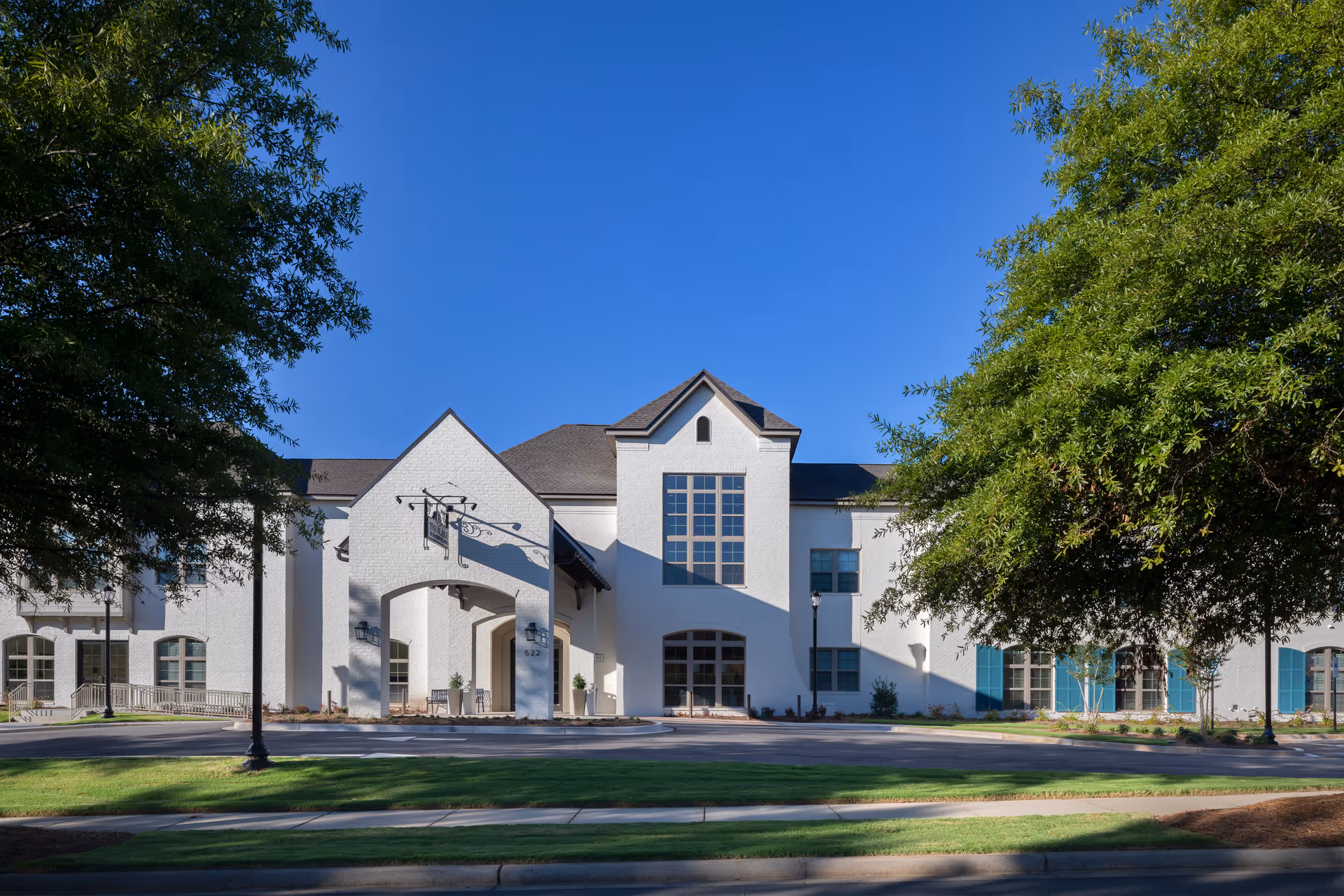 White two-story building with an arched entrance and large central window, framed by trees and a lawn.