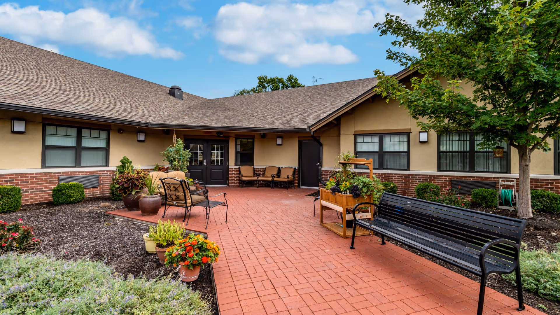 Outdoor courtyard area of a senior living facility with red brick paving, black metal bench, several potted plants with colorful flowers, cushioned chairs, and a tree. The building has beige walls with brick accents and multiple windows under a sloped roof. The sky is partly cloudy.