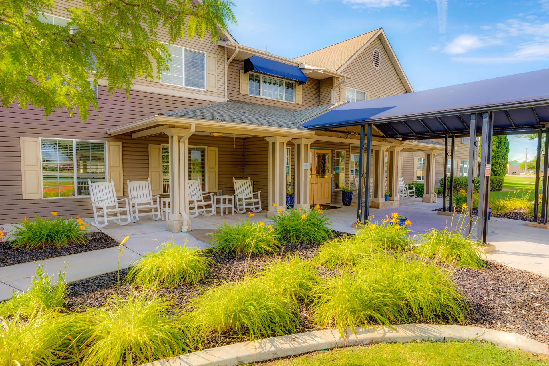 Exterior view of The Auberge at North Ogden senior living facility showing a covered entrance with a blue awning, beige siding, several white rocking chairs on the porch, and well-maintained landscaping with green bushes and flowers.