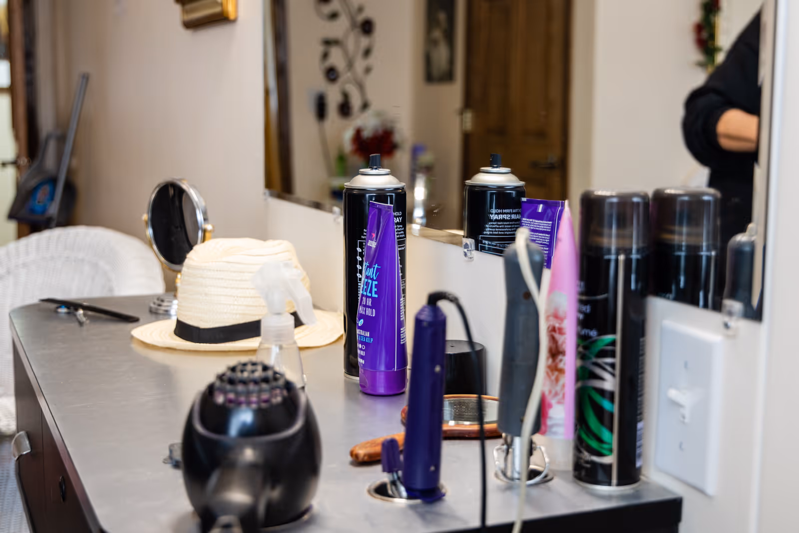 A countertop with various hair styling products including hairspray cans, a hair dryer, a spray bottle, a small round mirror, and a straw hat. A large mirror is mounted on the wall behind the counter reflecting some of the items and part of a person standing nearby.