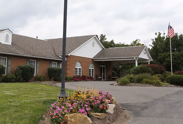 Exterior view of Mount Vernon of South Park facility showing a brick building with a covered entrance, surrounded by landscaped greenery, colorful flowers, and an American flag on a flagpole.
