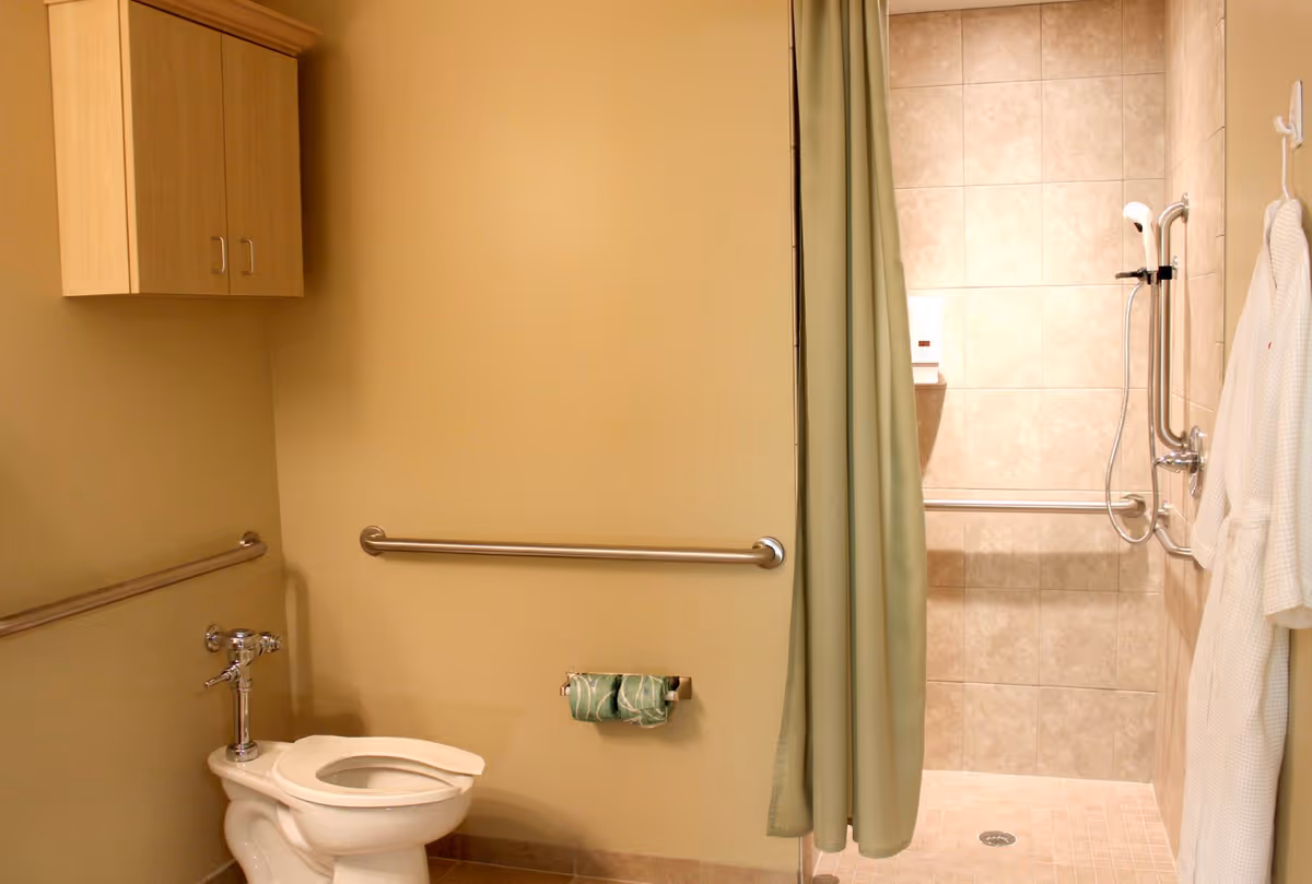 A bathroom with beige walls and tiled floor featuring a white toilet with a metal flush handle, two metal grab bars on the walls, a wooden cabinet mounted on the wall, a green shower curtain partially open revealing a tiled walk-in shower with a handheld showerhead and grab bars, and a white bathrobe hanging on a hook.