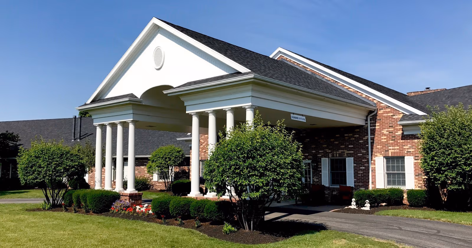 Columned white portico at the front entrance of a brick senior living building with landscaping and a driveway.