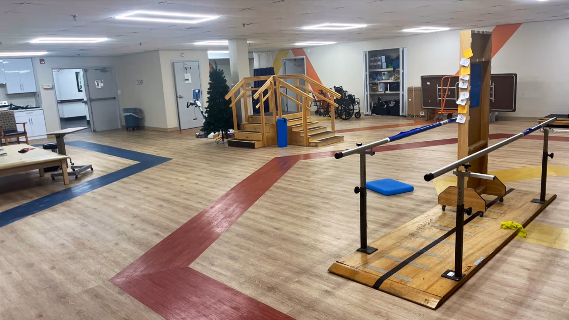 A spacious indoor rehabilitation or physical therapy room with wooden flooring marked by colored paths. The room contains parallel bars for walking exercises, a small set of wooden stairs with handrails, a blue cushion on the floor, and various mobility aids such as wheelchairs and walkers in the background. The walls are light-colored with some orange accents, and there is a Christmas tree near the center.