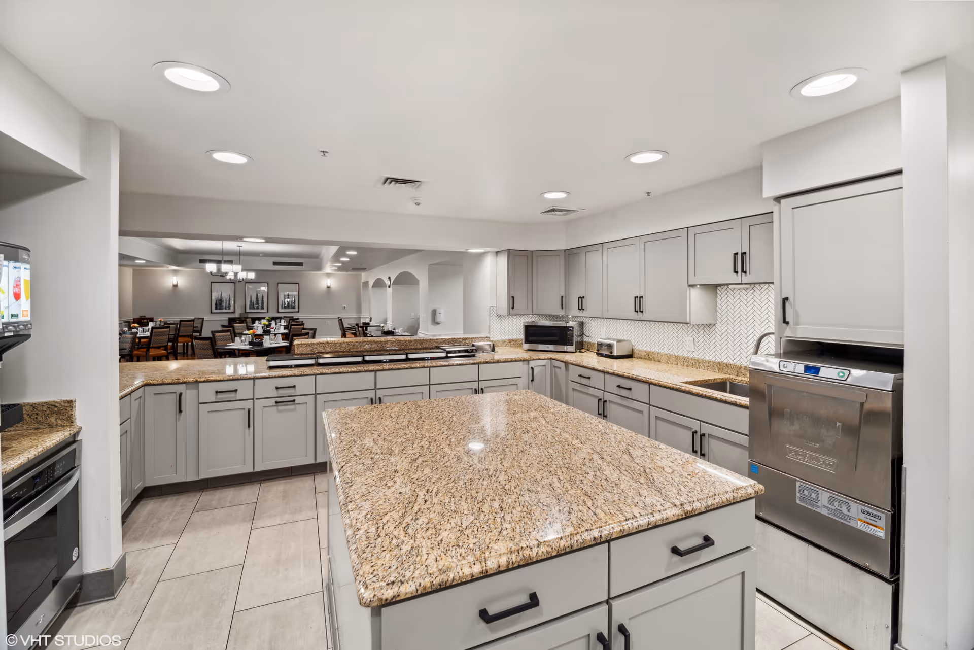 A modern kitchen area with granite countertops and gray cabinets. The kitchen features a large central island, a built-in oven, a microwave, a toaster, and a dishwasher. In the background, there is a dining area with tables and chairs, decorated with framed artwork on the walls and pendant lighting.