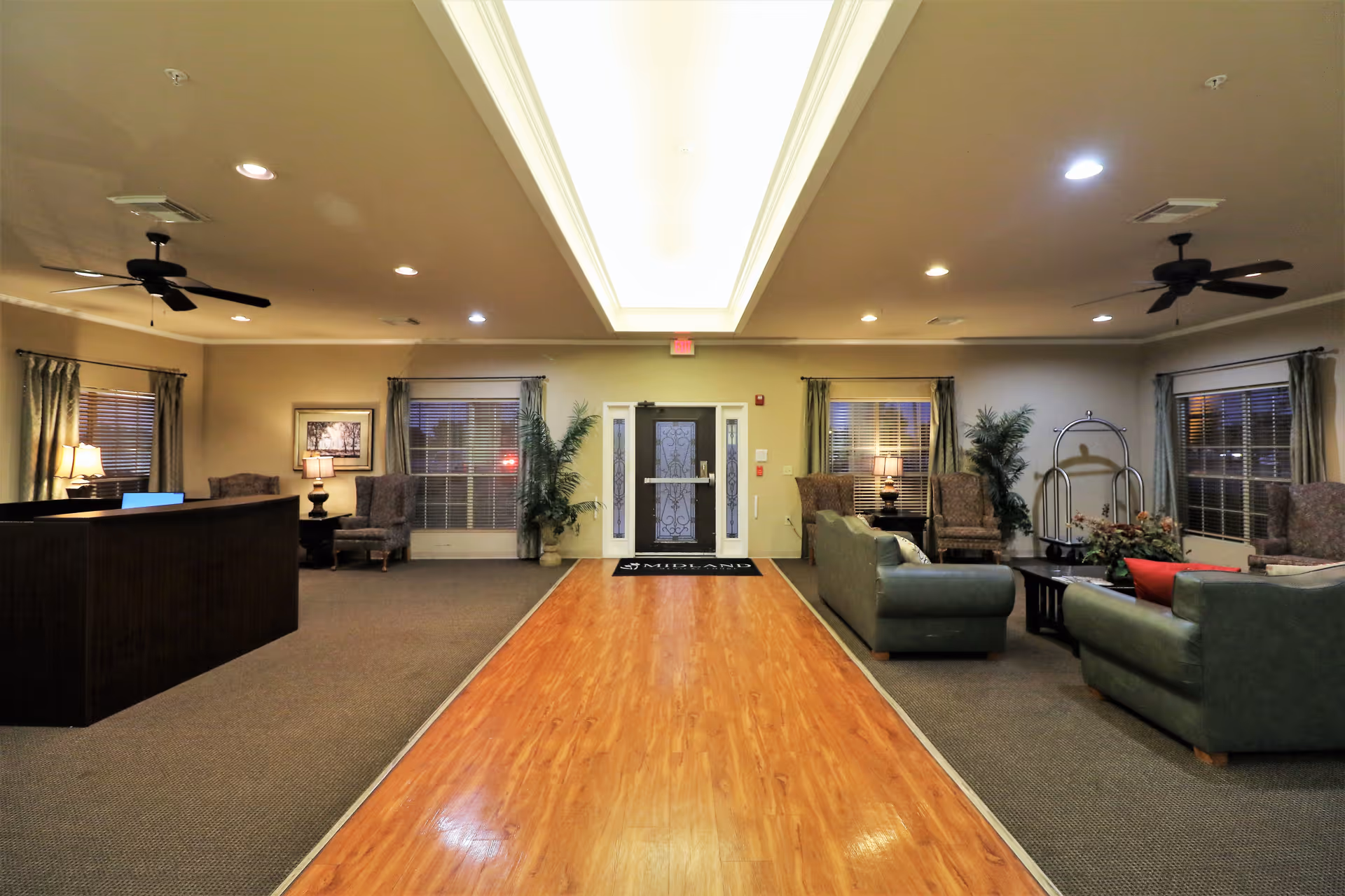 Interior view of a senior living facility lobby with a wooden floor pathway in the center, flanked by carpeted areas with green sofas, armchairs, side tables with lamps, and potted plants. Ceiling fans and recessed lighting are visible on the ceiling, and a glass door with sidelights is at the far end.