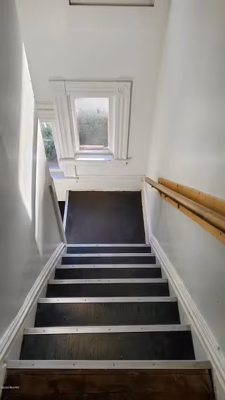A narrow indoor staircase with black steps and white trim, a wooden handrail on the right side, and a window with decorative molding at the top of the stairs letting in natural light.