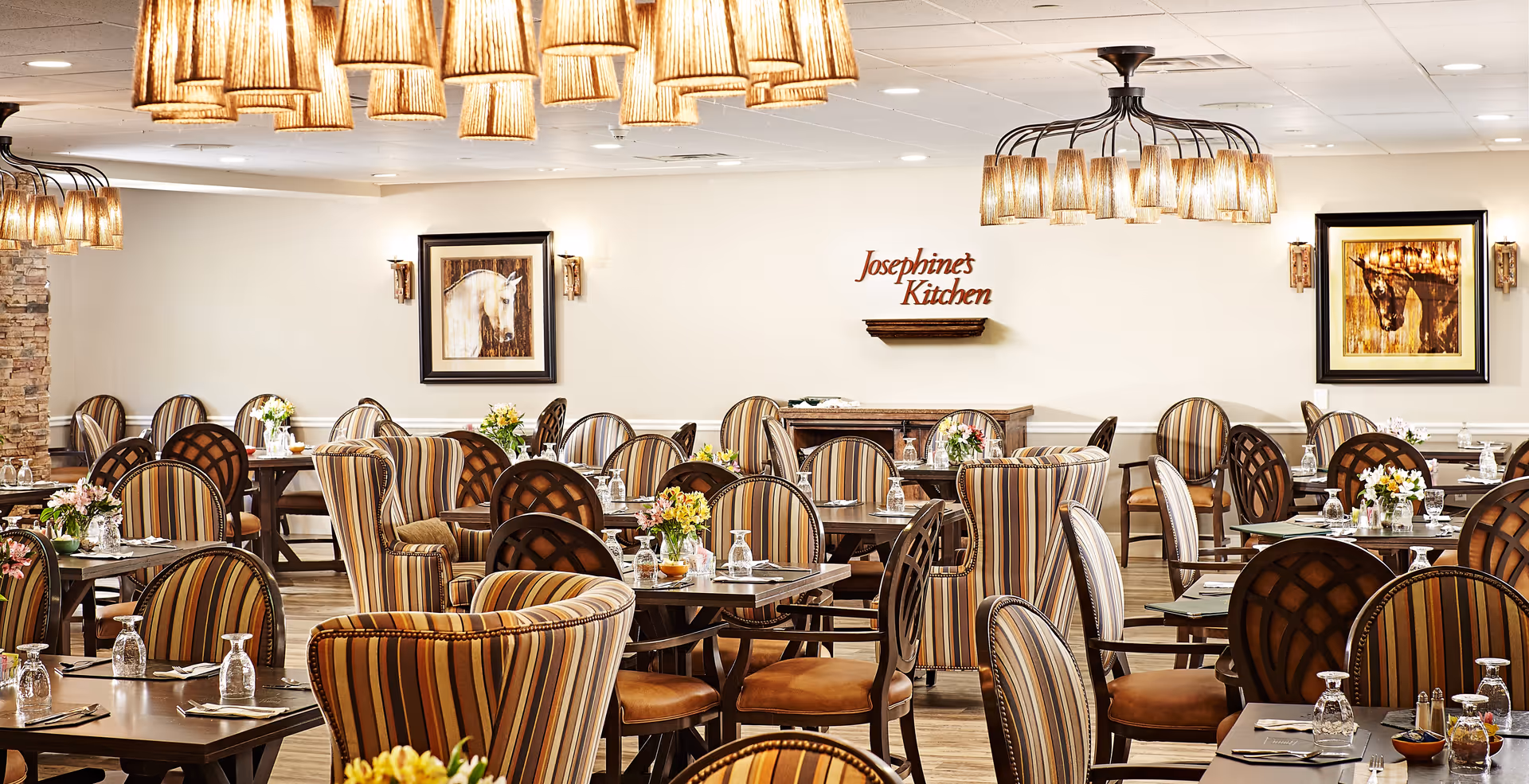 A spacious dining room with multiple tables and chairs arranged neatly. The chairs have striped upholstery in warm tones. The tables are set with glasses, napkins, and small flower arrangements. The room is well-lit with elegant hanging light fixtures. On the back wall, there are two framed paintings of horses and a sign that reads 'Josephine's Kitchen'.