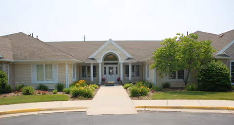 Front exterior view of a single-story building with beige siding and a brown shingled roof. The entrance features a covered porch with white columns and a large arched window above the door. There are landscaped flower beds with green shrubs and yellow flowers on either side of the walkway leading to the entrance.