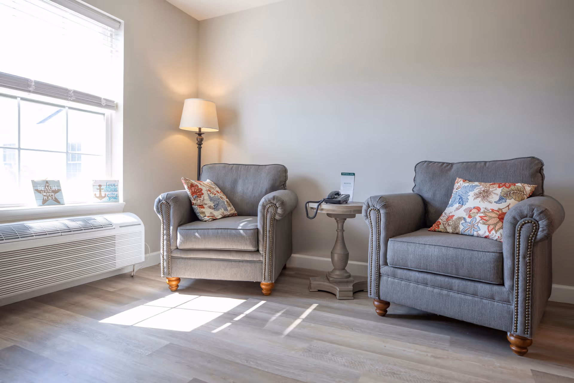 A cozy sitting area with two gray armchairs featuring floral patterned cushions, a small round wooden side table with a telephone and a card, a floor lamp in the corner, and a large window with blinds letting in natural light. The floor is light wood and the walls are painted a soft neutral color.
