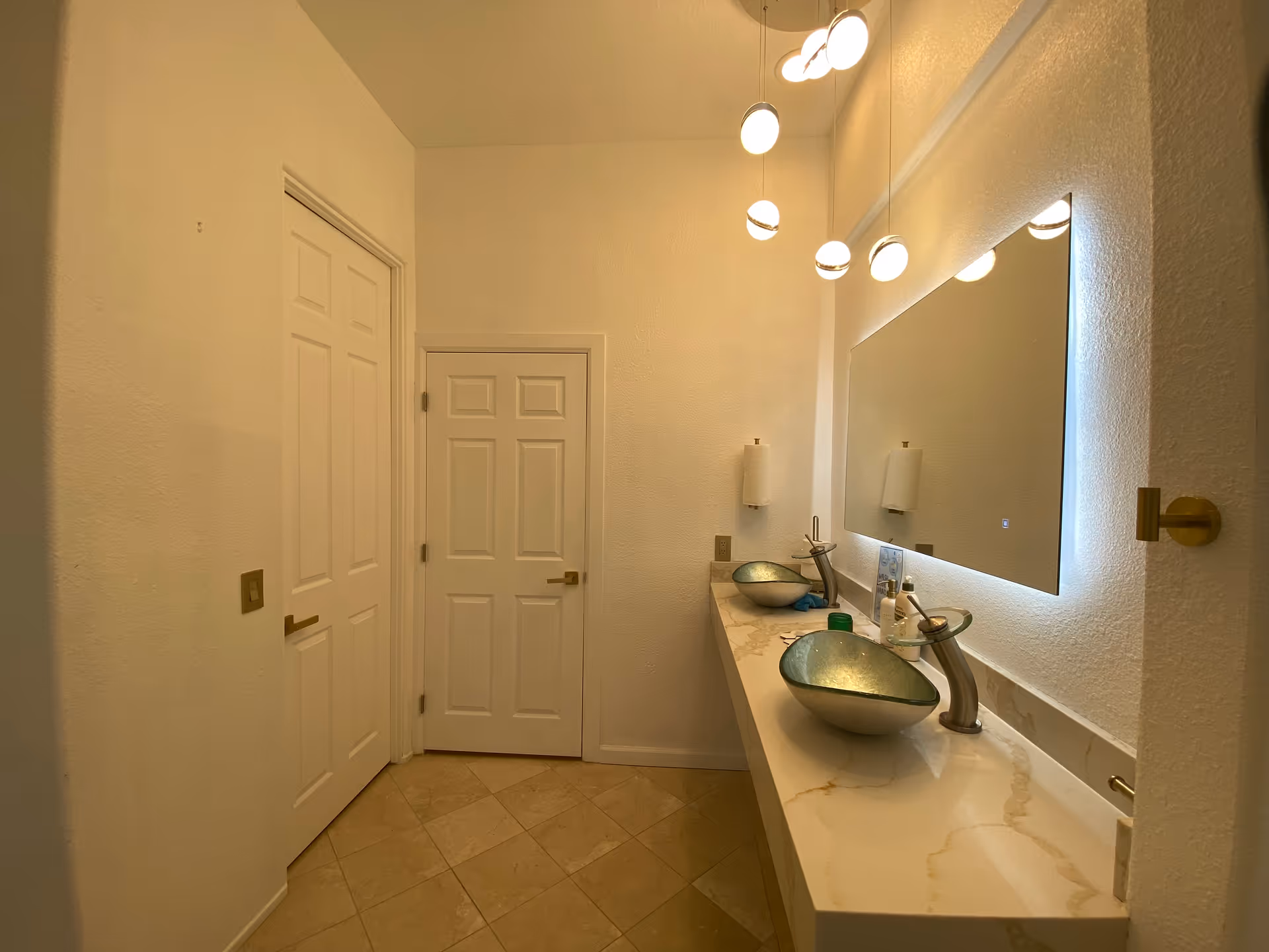 Well-lit bathroom with two vessel sinks on a marble countertop, an illuminated mirror, pendant lights, and two white doors.