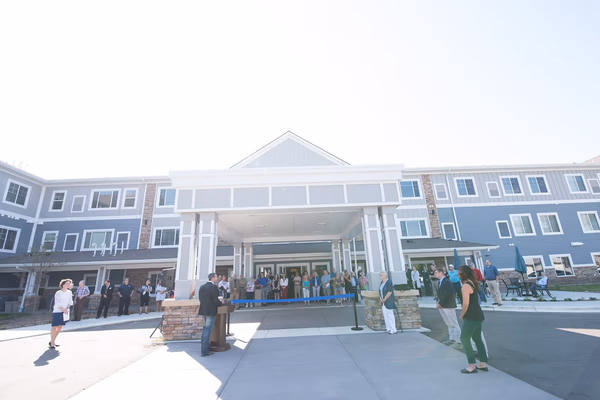 A group of people gathered outside the entrance of a large, modern senior living facility named Havenwood of Richfield. The building has a covered drop-off area with white pillars and a blue-gray exterior with multiple windows. Some people are standing near a podium, possibly for a speech or ribbon-cutting event.