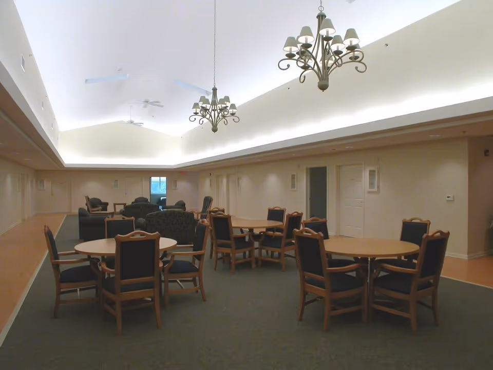 A spacious common area with round wooden tables and chairs arranged on a carpeted floor. In the background, there are several upholstered armchairs and sofas. The room has a high ceiling with two decorative chandeliers and ceiling fans, and the walls are painted a light color with several doors visible.