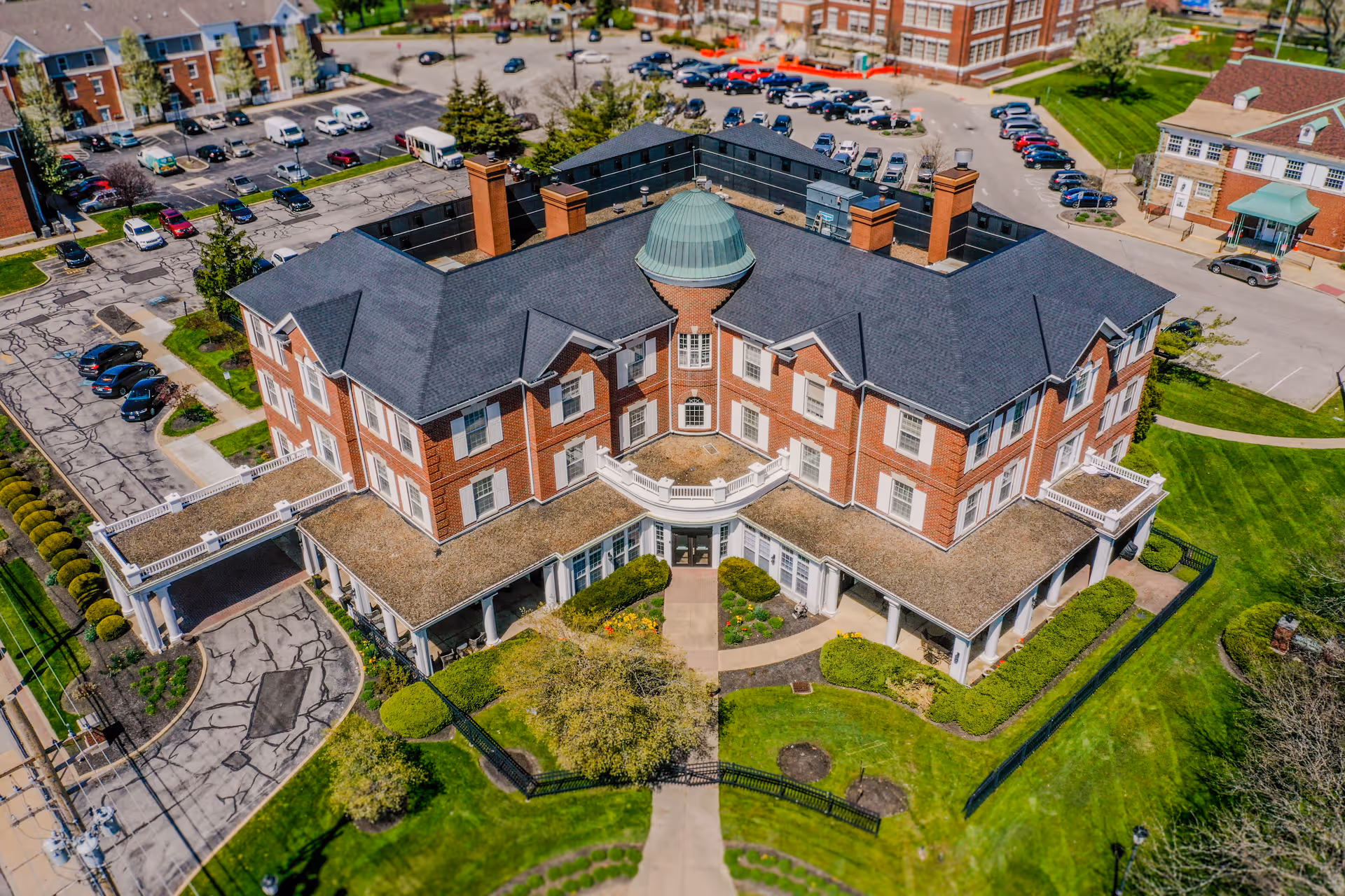 Aerial view of a large, multi-story brick building with a dark shingled roof and a central dome. The building is surrounded by well-maintained green lawns, shrubs, and a paved driveway leading to a covered entrance. Several cars are parked in the parking lot adjacent to the building, and other buildings are visible in the background.