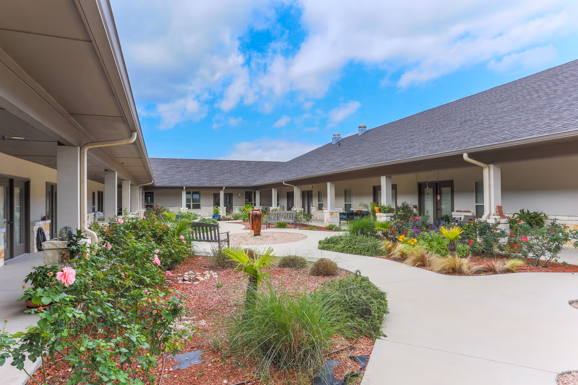 Outdoor courtyard area of Arbor House Assisted Living and Memory Care featuring a paved walkway, garden beds with various plants and flowers, benches, and a water fountain under a partly cloudy sky.