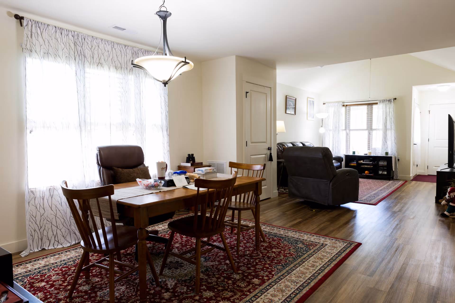 Open-plan dining and living area with a wooden dining table and chairs on a patterned rug, reclining chairs and a TV near large windows with sheer curtains.