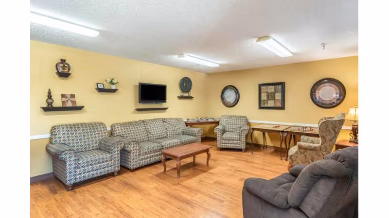 A cozy living room area in a nursing and rehab facility with patterned armchairs and a sofa arranged around a wooden coffee table. The walls are painted light yellow and decorated with framed artwork and small shelves holding decorative items. A flat-screen TV is mounted on the wall above a shelf. The floor is wood, and there are tables along the far wall with board games on them.