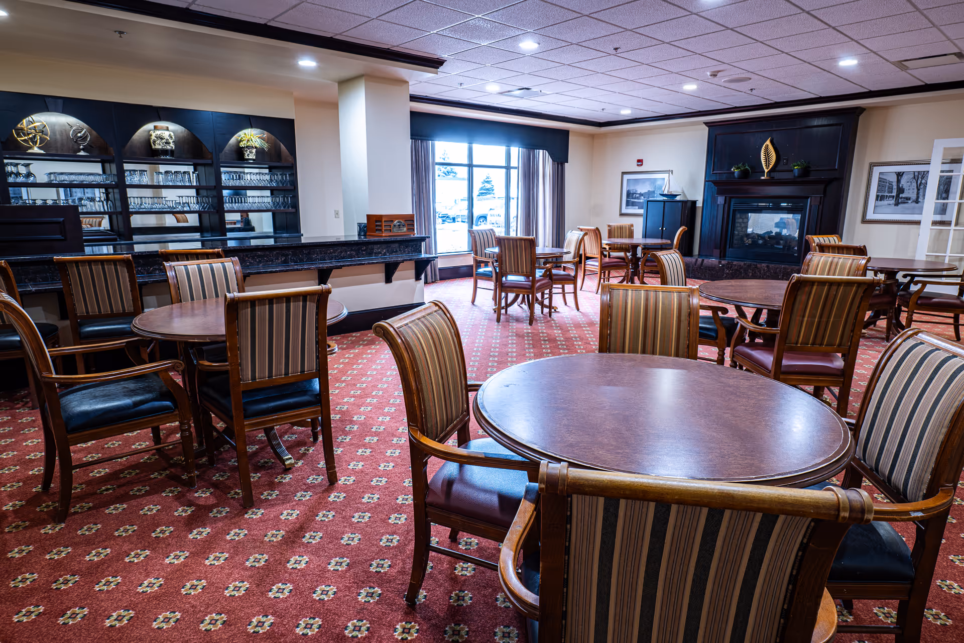 A spacious dining room with multiple round wooden tables and striped upholstered chairs arranged neatly on a red patterned carpet. The room features a large window with curtains, a dark wooden fireplace with decorative items on the mantel, and a built-in cabinet with glassware and decorative pieces. The ceiling has recessed lighting and a grid pattern.