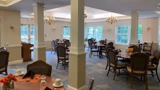 A spacious dining room with multiple round tables and chairs arranged neatly. The room features large windows allowing natural light to enter, beige walls, and chandeliers hanging from the ceiling. Tables are set with cups, plates, and small flower arrangements.