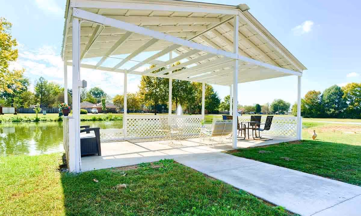 A white open-air pavilion with a pitched roof situated next to a small pond. The pavilion contains outdoor seating including chairs and tables, surrounded by green grass and trees under a clear blue sky.