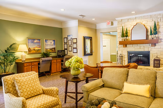 A cozy living room area in an assisted living facility featuring a beige sofa with a yellow pillow, a patterned armchair, a wooden coffee table with a green vase of flowers, and a stone fireplace with decorative plants and a mirror above the mantle. In the background, there is a workspace with two desks, chairs, a lamp, framed pictures, and certificates on the wall.