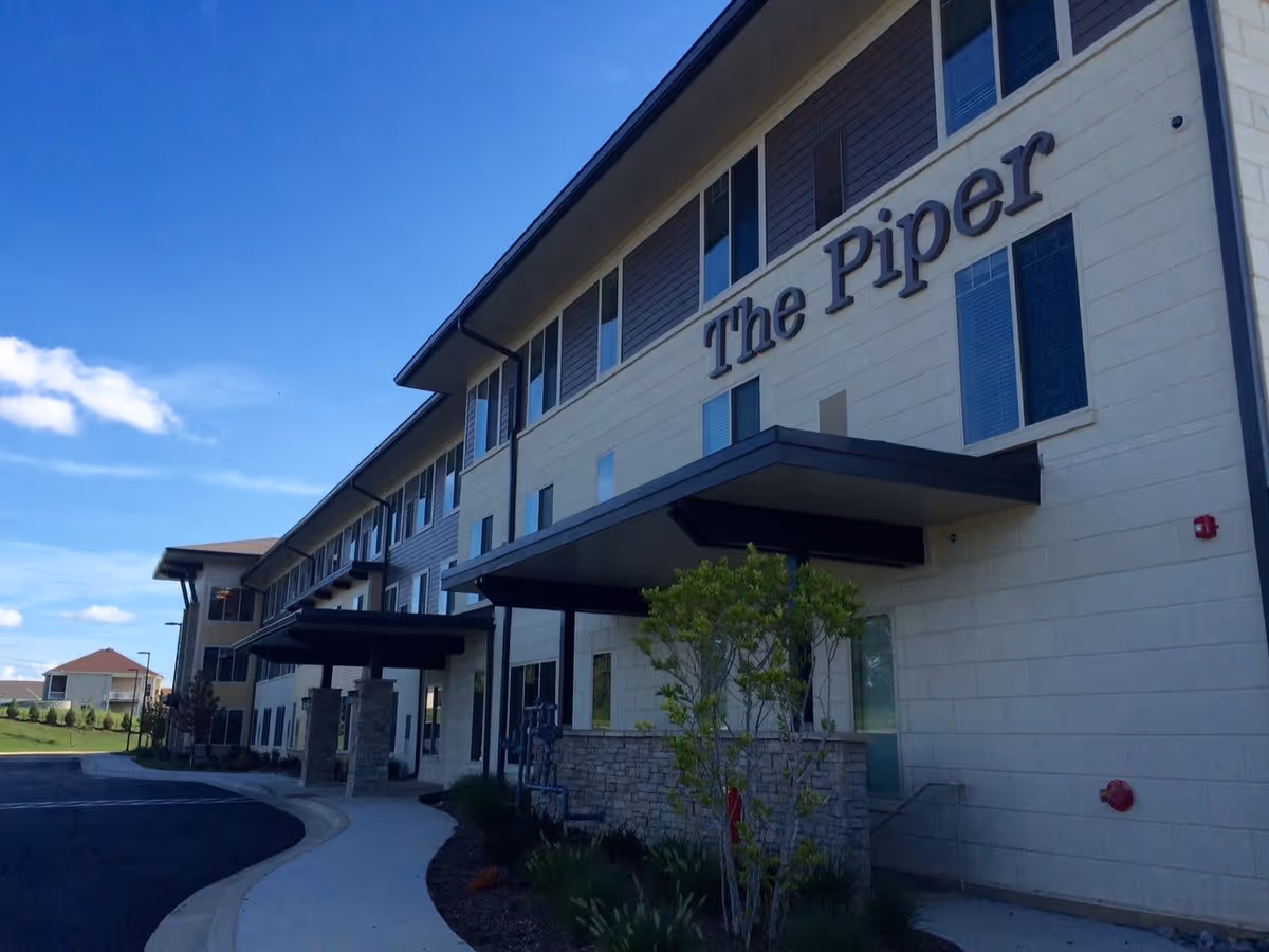 Exterior view of The Piper Assisted Living and Memory Care building on a clear day, showing a multi-story structure with windows, a covered entrance, and a curved sidewalk leading to the entrance.