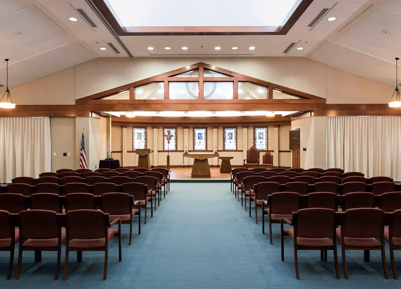 Interior view of a chapel or worship room with rows of brown chairs facing a raised platform with a wooden altar, lecterns, and stained glass windows in the background. The ceiling has wooden beams and recessed lighting, and there are curtains on the side walls.