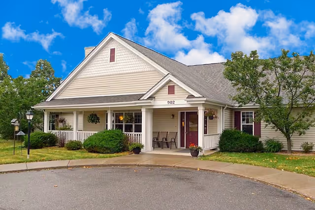 Exterior view of a single-story senior living facility building with beige siding, white trim, and a gray shingled roof. The entrance has a covered porch with white columns, two chairs, and potted plants. The building number 902 is visible above the entrance. There are bushes and a tree around the building, with a clear blue sky and some clouds in the background.