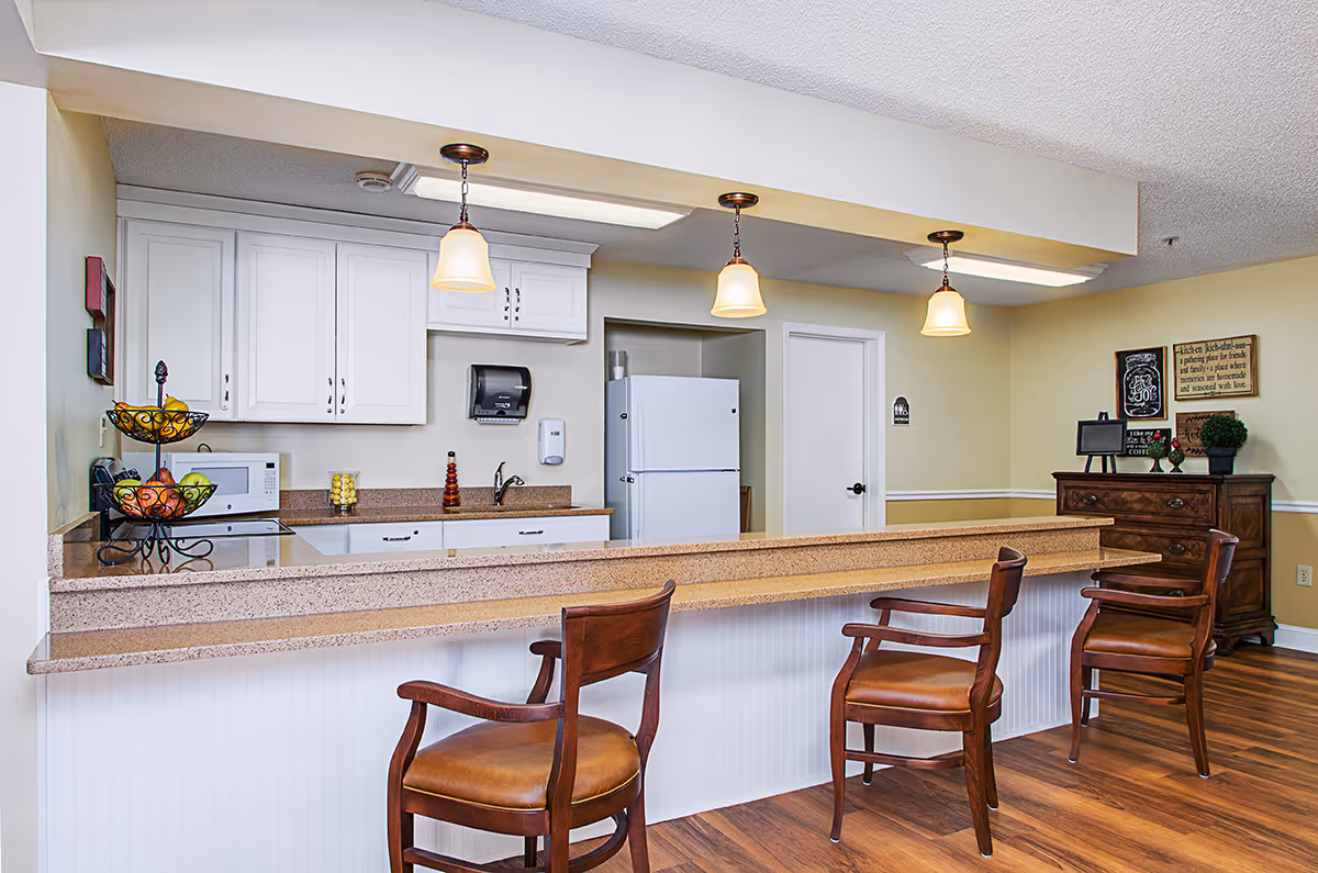 A clean and well-lit kitchen area with a long beige countertop and three wooden chairs with brown leather seats. The kitchen has white cabinets, a white refrigerator, a microwave, and a paper towel dispenser. Three pendant lights hang above the counter. In the background, there is a wooden dresser with decorative items and framed signs on the wall.