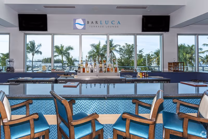 Interior view of Bar Luca Terrace Lounge with a bar counter, several bottles of liquor displayed behind the counter, and four wooden bar stools with blue cushions in front. Large windows behind the bar show palm trees and a marina outside.