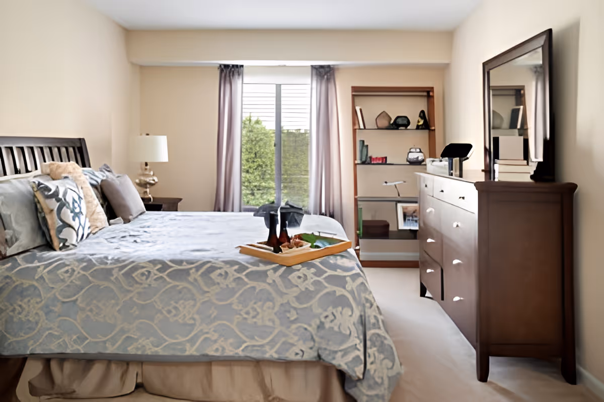 A neatly made bed with patterned blue and beige bedding and multiple pillows in a bedroom. A wooden dresser with a large mirror and various items on top is on the right side. A wooden bookshelf with decorative items and books is in the background near a window with light purple curtains. A tray with a bottle and food is placed on the bed.