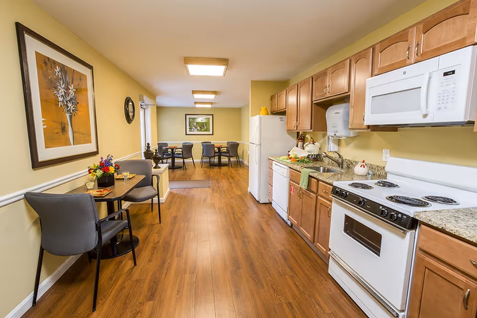 A bright kitchen and dining area in a senior living facility featuring wooden cabinets, white appliances including a stove, microwave, refrigerator, and dishwasher. There are two small dining tables with chairs, one near the wall with a flower arrangement and the other at the far end of the room. The walls are painted light yellow and decorated with framed artwork. The floor is wooden.