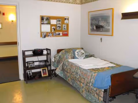 Bedroom in Newark Manor Nursing Home with bed, quilt, bulletin board, and framed photos.