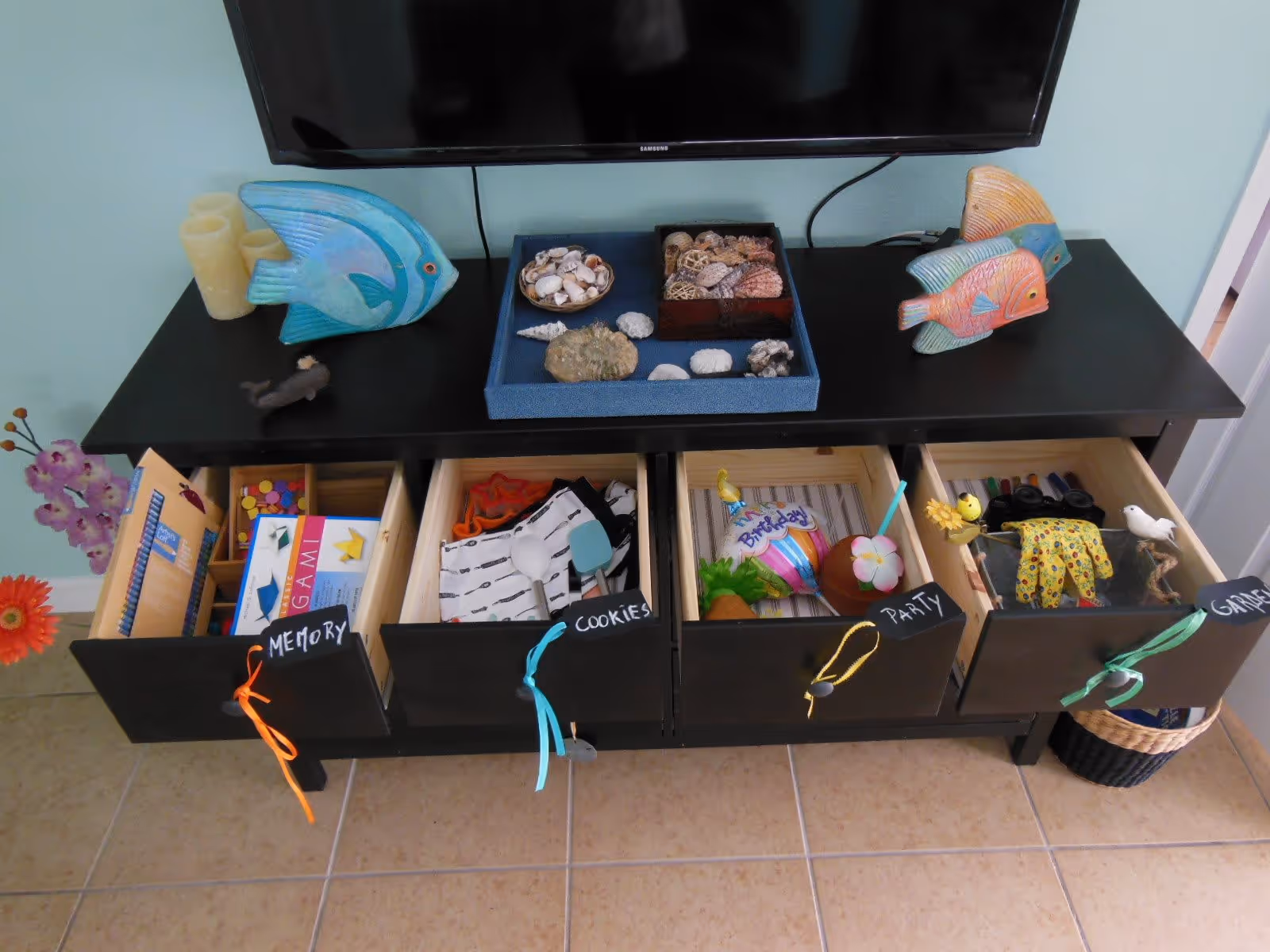 A black cabinet with four open drawers labeled Memory, Cookies, Party, and Garden, containing various items such as books, games, decorations, and craft supplies. On top of the cabinet are decorative fish sculptures, candles, and trays filled with seashells. A flat-screen TV is mounted on the wall above the cabinet.