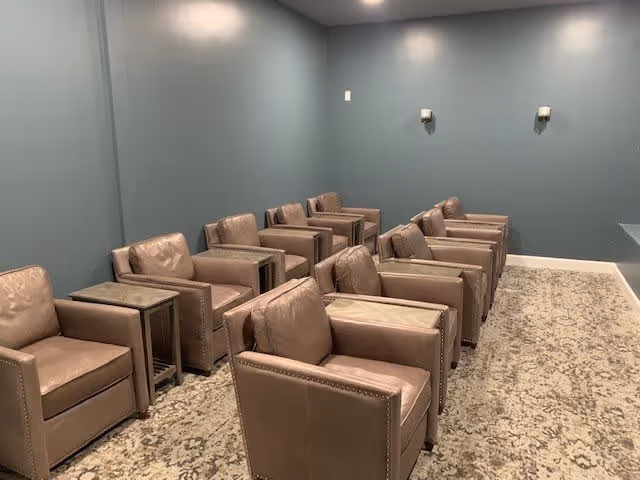 A room with two rows of beige leather armchairs separated by small side tables, set against blue-gray walls with two wall-mounted light fixtures and a patterned carpeted floor.