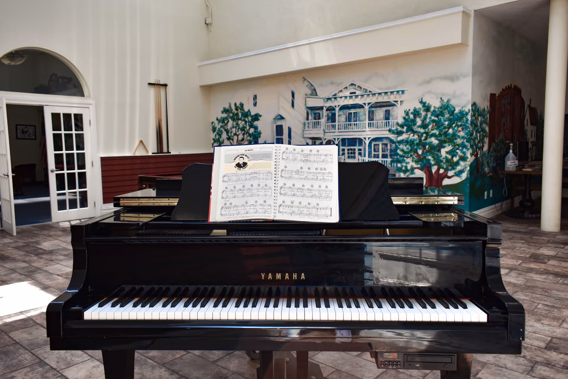 A black Yamaha grand piano with sheet music displayed on the music stand, situated in a spacious room with tiled flooring. The background features a mural of a large house with trees and an open doorway leading to another room.